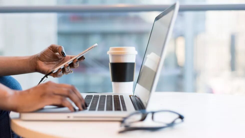 Aufgeklappter Laptop. eine Hand an der Tastatur eine hält ein Smartphone. Im Hintergrund steht ein Kaffeebecher, im Vordergrund eine zusammengeklappte Brille.