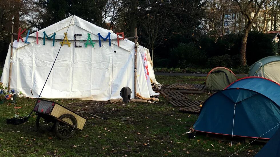 Man sieht ein großes weißes Veranstaltungszelt, auf dem in bunten Buchstaben steht: Klimacamp. Daneben stehen mehrere kleinere Camping-Zelte.