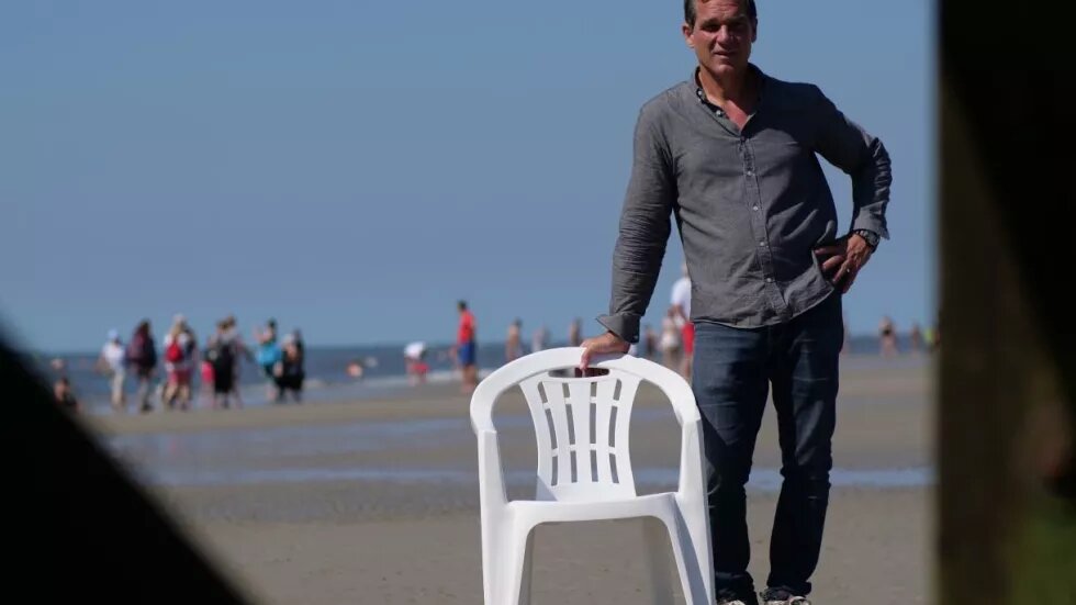 Der Regisseur des Films Monobloc, Hauke Wendler, steht am Strand und stützt sich auf einen weißen Plastikstuhl.