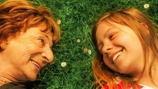 Eine ältere Frau und ein Mädchen liegen auf einer Wiese, ihre Köpfe liegen dicht beieinander und sie sehen sich lachend an.